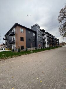 A modern, four-story apartment building labeled "MIDTOWN" stands on a quiet, empty street, with balconies on each floor and some trees with autumn leaves lining the sidewalk under a cloudy sky.