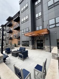 Modern apartment building with gray and brown exterior, orange awning labeled "415," and an outdoor seating area with blue cushioned chairs, tables, and a fire pit on a concrete patio.