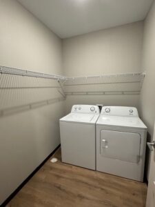 A laundry room with a washing machine and dryer side by side under a long white wire shelf; the walls are light beige and the floor is wood-style.