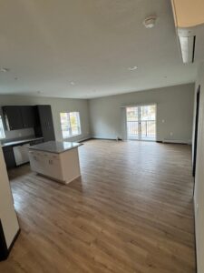 Spacious, unfurnished open-plan apartment with light wood flooring, a modern kitchen with dark cabinets and a white island, large windows, and sliding glass doors leading to a balcony.