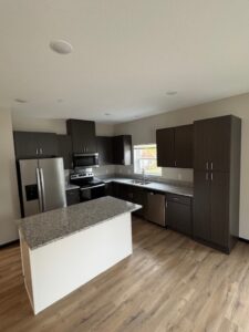 Modern kitchen with dark wood cabinets, granite countertops, a central island, stainless steel appliances, and light wood flooring. There is a window above the sink letting in natural light.
