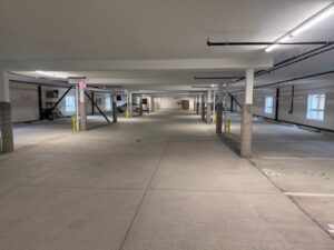 A clean, mostly empty indoor parking garage with a few vehicles parked along the left side. Concrete columns and beams support the ceiling, and fluorescent lights illuminate the space.