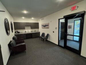 A small waiting area with two dark chairs, a kitchenette with cabinets and sink, two chairs against the wall, a large glass exit door, and circular mirrors on the wall. The space is clean and modern.