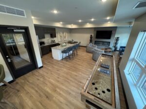 Modern lounge with wood flooring, a shuffleboard table, kitchen area with barstools, gray sofas, large TV, and windows letting in natural light. Neutral decor with wall art and decorative sculptures.