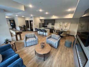 A modern lounge with a kitchenette, seating area, and game tables. The room has wood flooring, blue and patterned chairs, a round coffee table, bar stools, and wall art with large leaf prints.