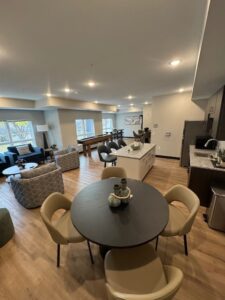 Modern open-concept living space with wood flooring, round dining table, kitchen island, lounge area with sofas and chairs, and large windows providing natural light. Neutral-colored decor and stylish lighting throughout.